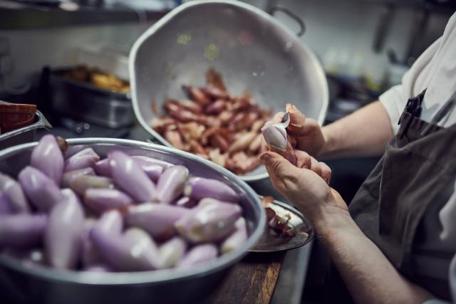 A chef is seen preparing ingredients. A bowl of peeled shallots is visible in the foreground.