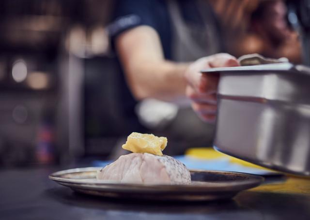 a beautifully plated dish is shown in the foreground. A chef is moving a gastronorm pan.